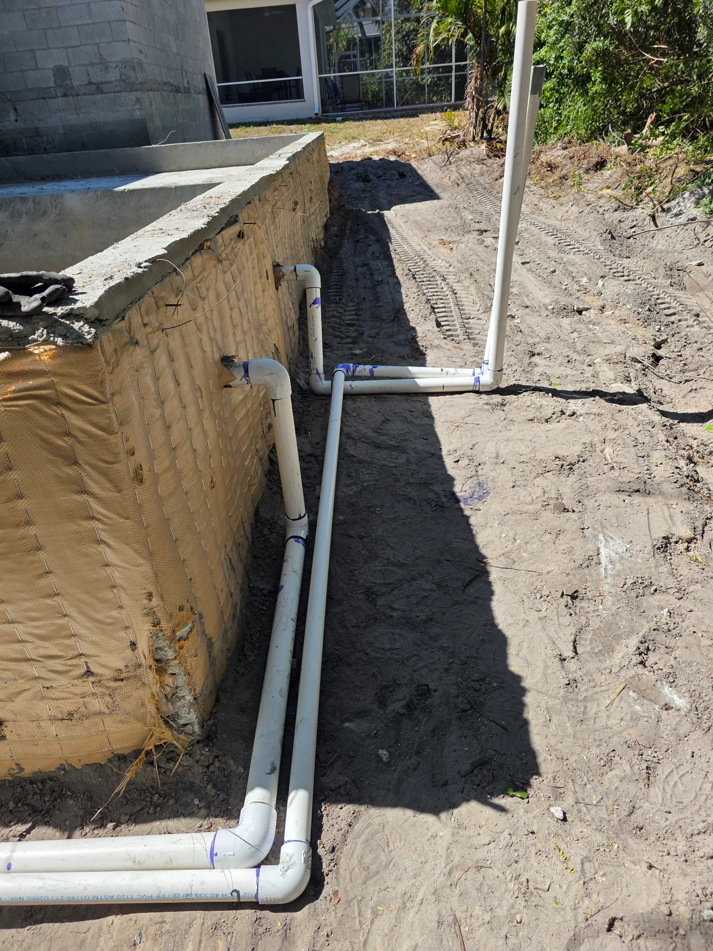 PVC drainage pipe installation in trench next to pool equipment area with concrete foundation
