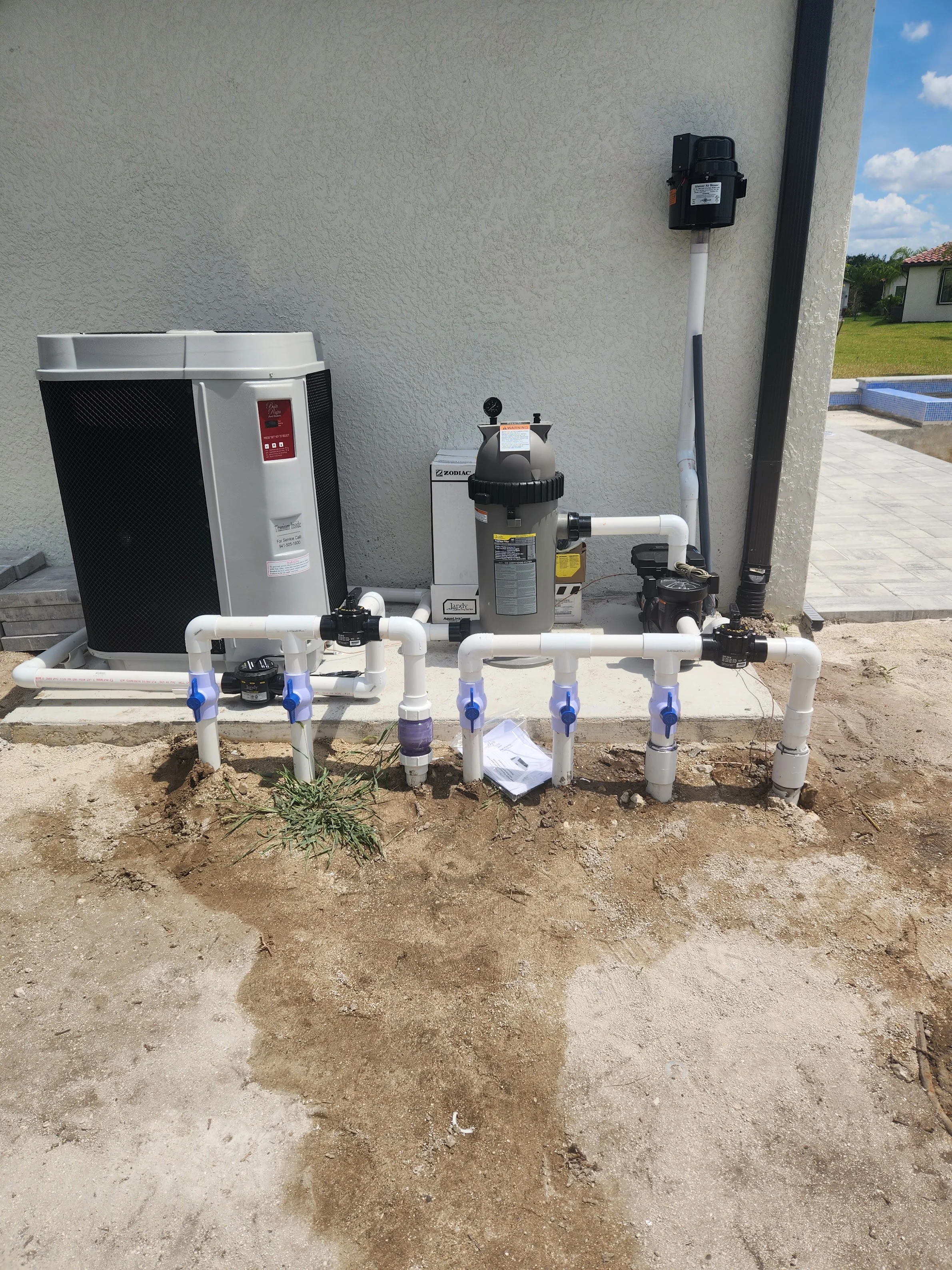 A pool technician inspecting the plumbing connections at a residential pool equipment pad in Lee County, Florida. PVC return and suction lines are visible alongside the pump and filter system during a repair assessment.